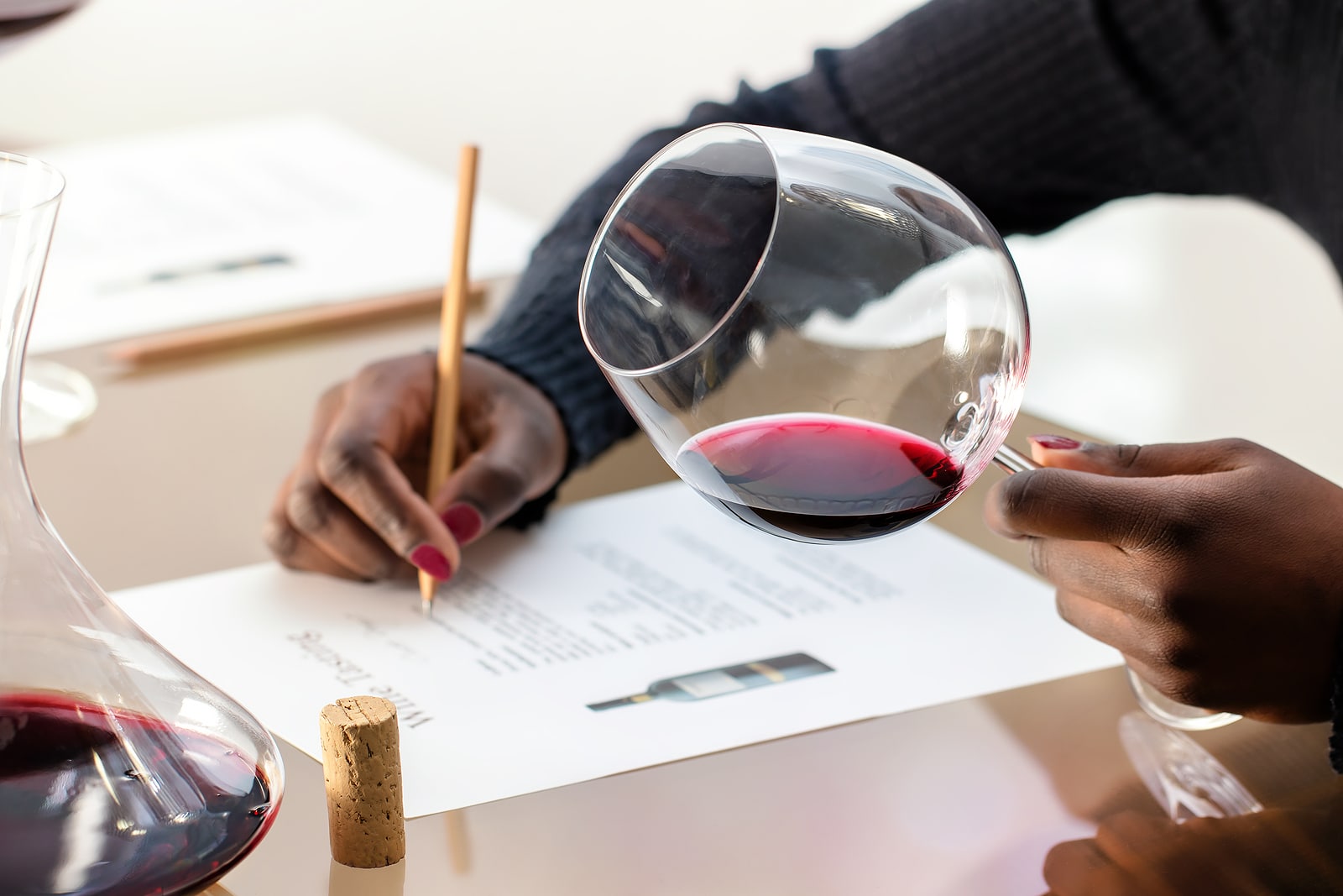 Close up detail of hands holding red wine glass.Wine enthusiast evaluating red wine at wine tasting. Woman taking notes with decanter at table.