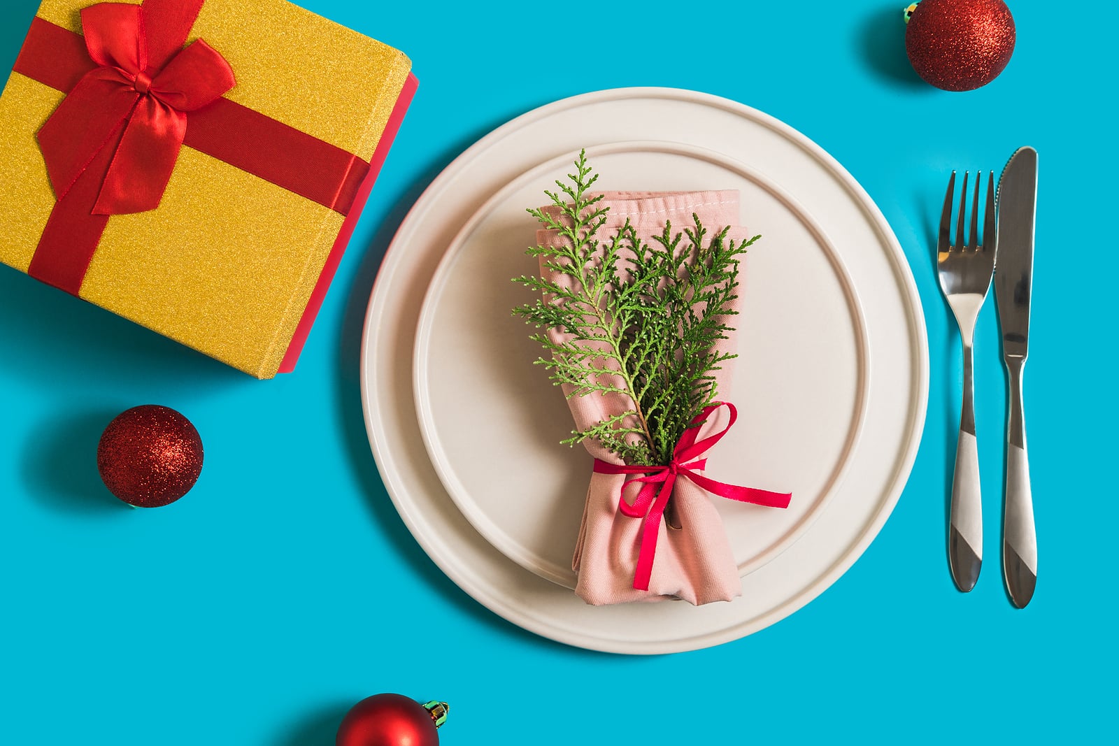 Served table with plate and cutlery for celebration of Christmas and New Year. On plate is napkin with a Christmas tree branch, red balls. Flatlay on blue background with balls, gift box.
