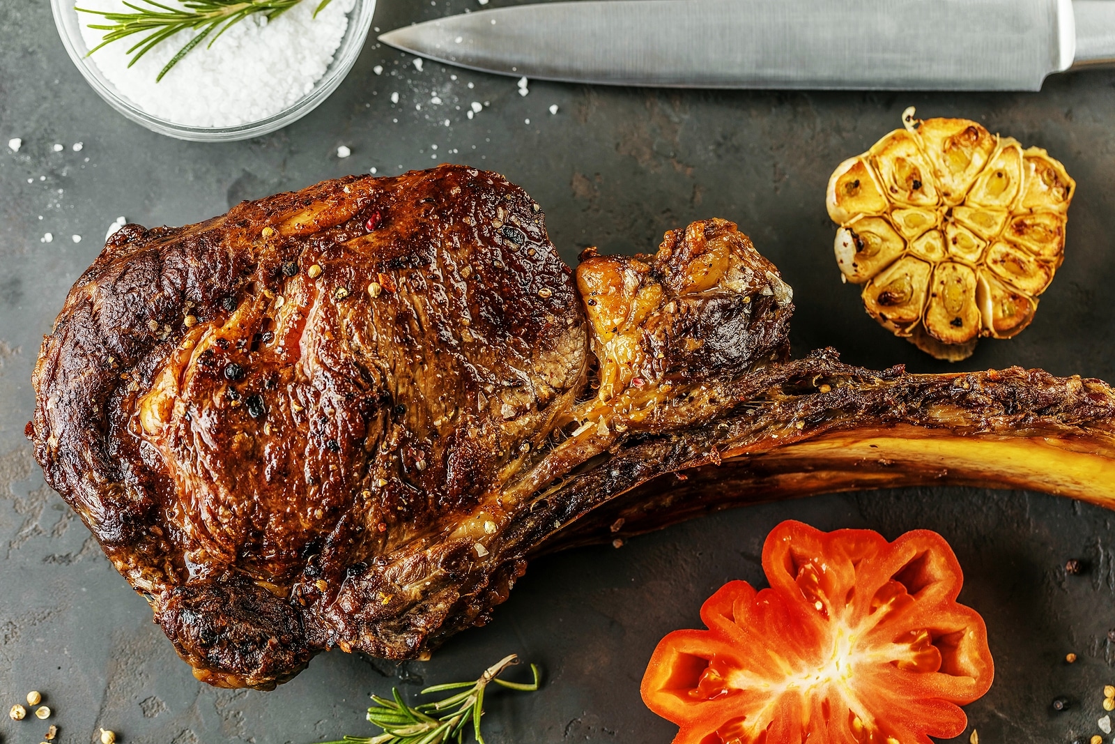 Sliced rare grilled or barbecued tomahawk beef steak on a griddle with fresh rosemary and tomato in a close up view.